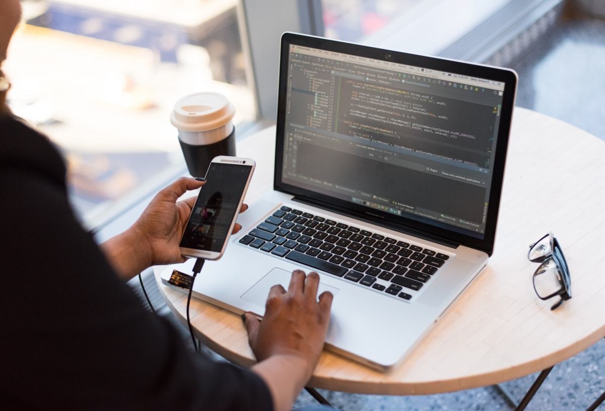 Person working on laptop with code editor open, holding a smartphone, with a coffee cup on the table.