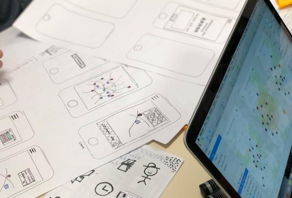 Sketches of mobile app screens on paper next to a laptop displaying a map.